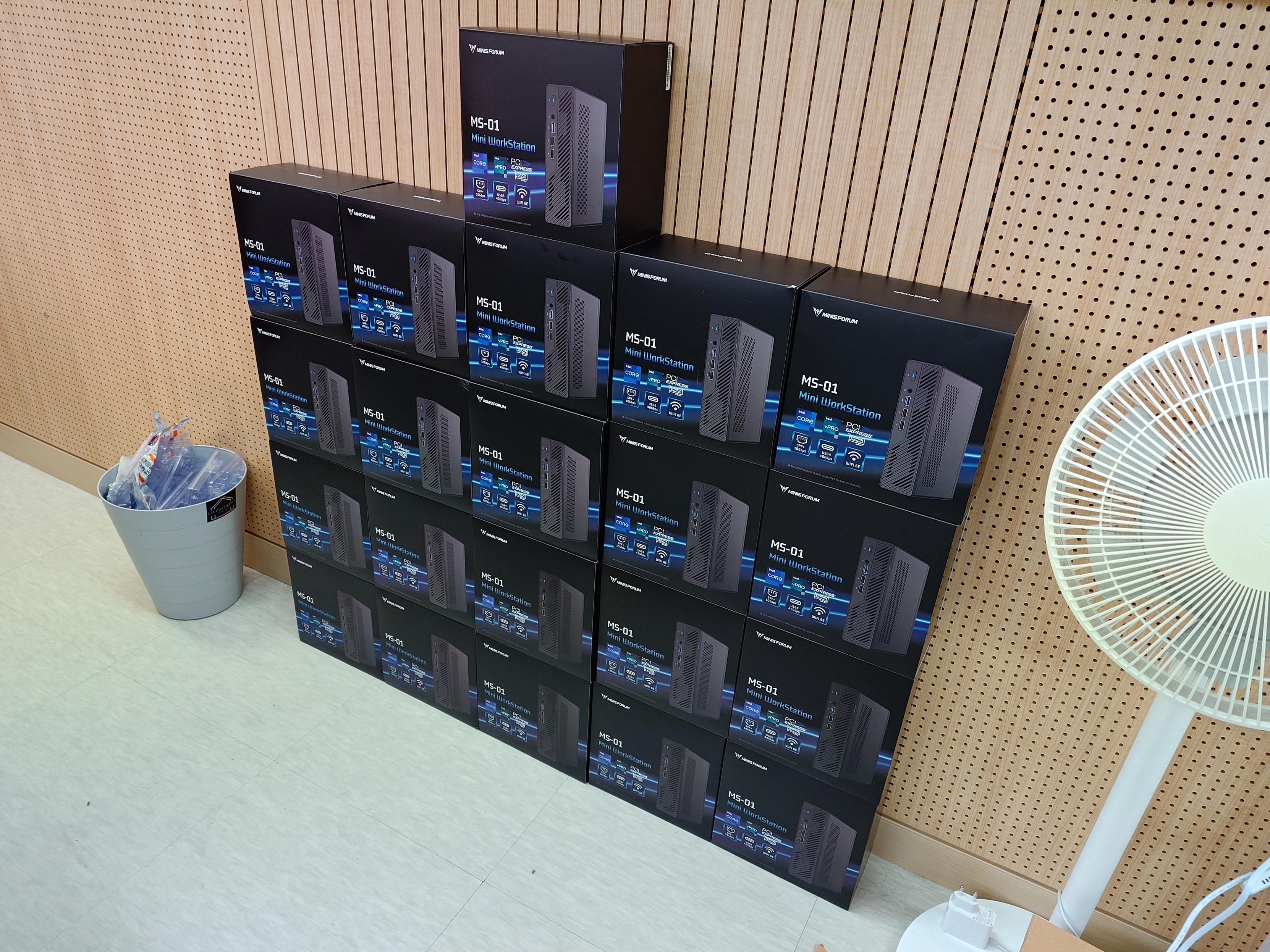 Minisforum MS-01 제품 상자 여러 개를 벽 쪽에 높게 쌓아 둔 사진.